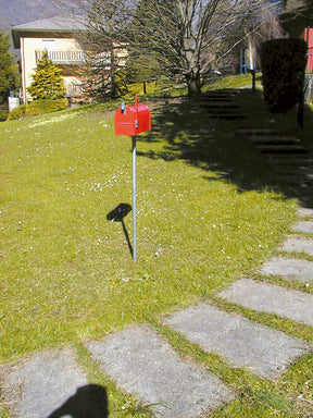 Cassetta delle lettere rossa su palo metallico in un giardino con prato e vialetto in pietra.